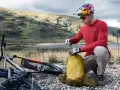 Kolarz w kasku przygotowuje żółty plecak Deuter, idealny jako jaki plecak na rower, obok leżącego roweru w malowniczym krajobrazie.