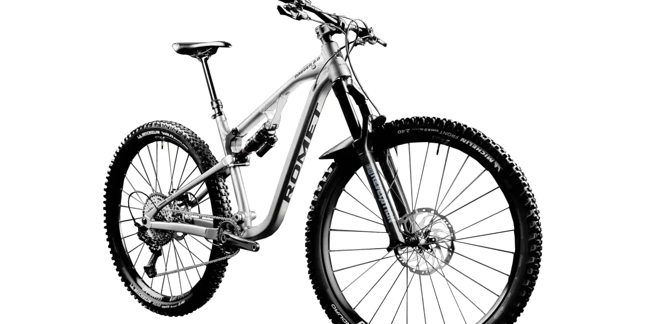 Srebrny rower górski Romet Dagger 2.0 z pełnym zawieszeniem, oponami Michelin Wild Enduro i obręczami DT Swiss M1700. Jedna z wielu marek rowerów.