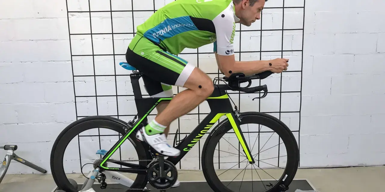 Mężczyzna w stroju kolarskim podczas profesjonalnego fittingu roweru. Czarny rower z zielonymi akcentami stoi na trenażerze.