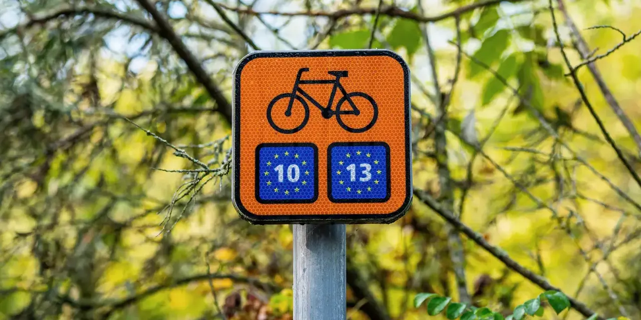 Pomarańczowy znak z rowerem i unijnymi flagami z numerami 10 i 13, wskazujący trasę Euro Velo.