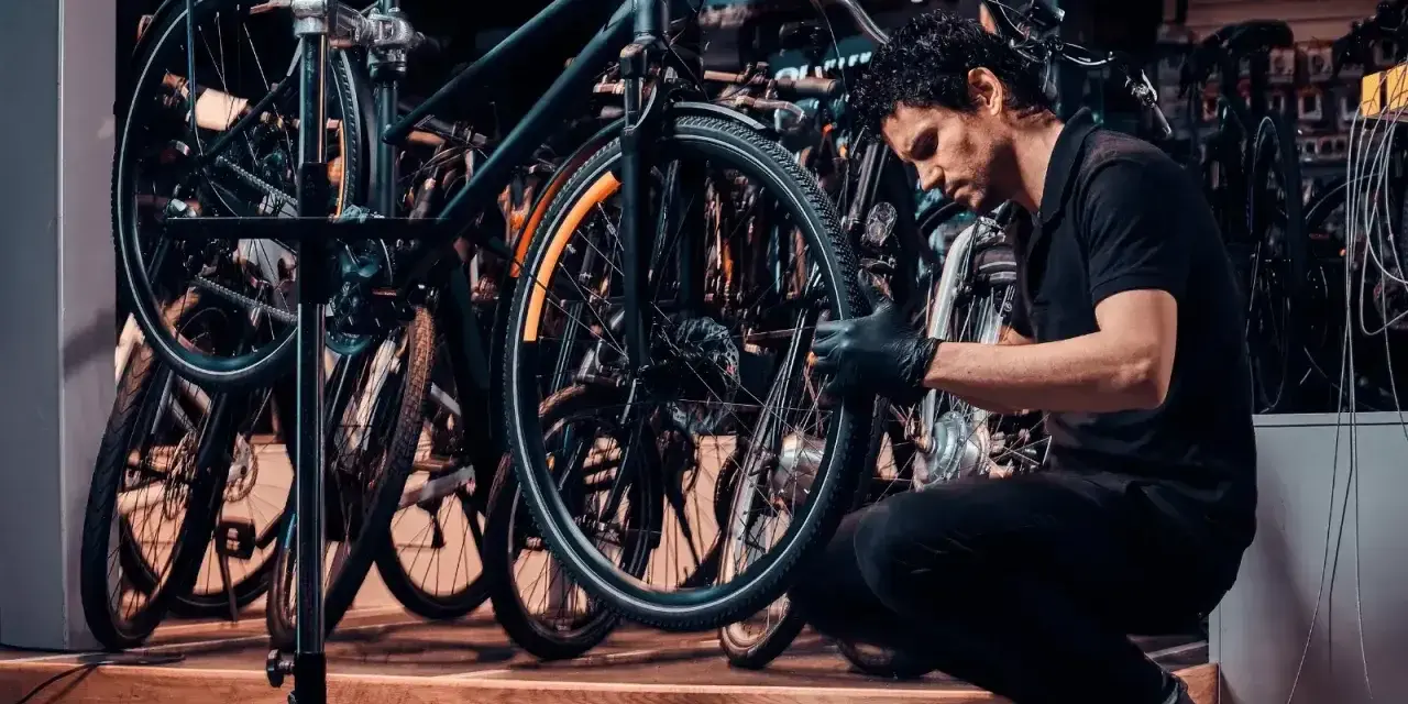 Mechanik w czarnej koszulce i rękawiczkach pokazuje, jak założyć tylne koło w rowerze, pracując w serwisie rowerowym.