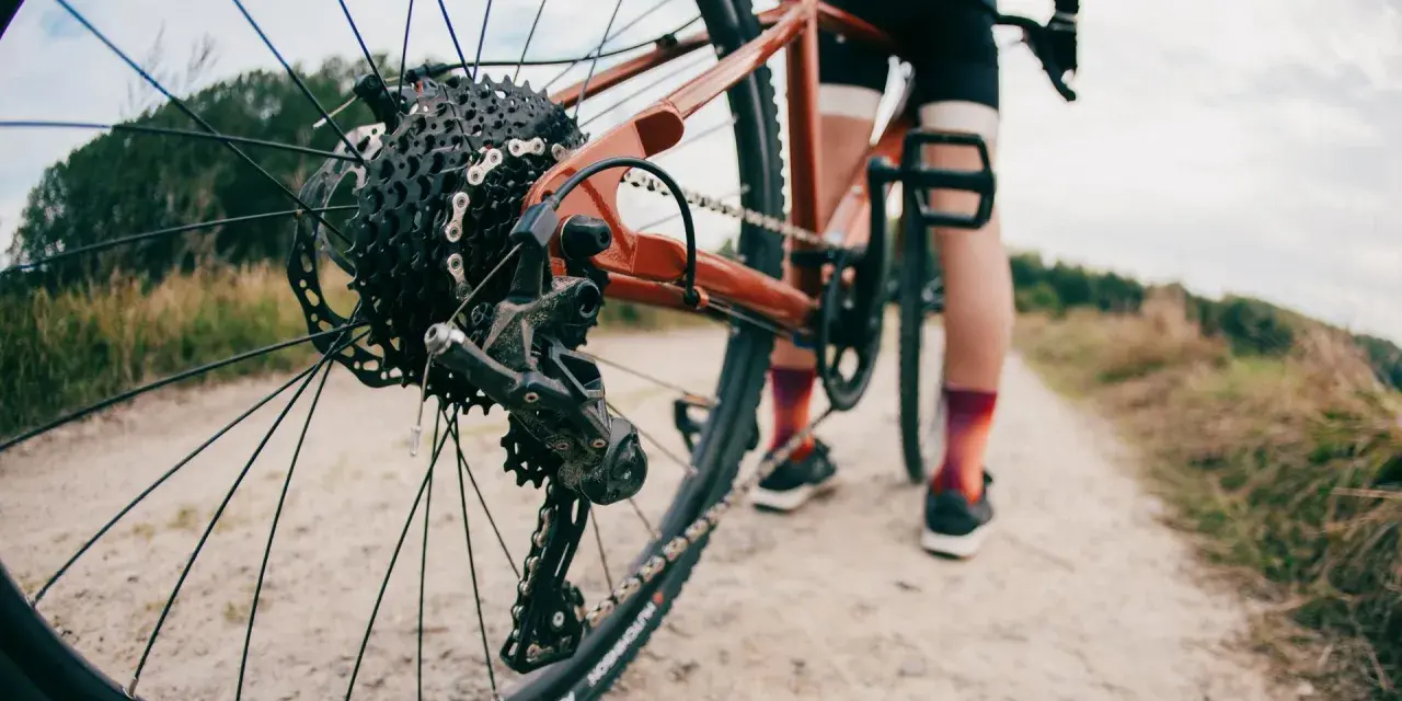 Zbliżenie na tylną przerzutkę roweru gravelowego, gdzie widać sprzęgło w przerzutce. Rower stoi na szutrowej drodze, a w tle widać sylwetkę rowerzysty.