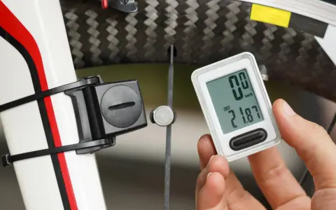 Dłoń trzyma licznik rowerowy pokazujący dystans 21.87 km. Obok widoczny element czujnika na obwodzie koła rowerowego.