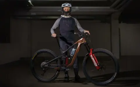 Kolarz w kasku i odzieży rowerowej prezentuje swój nowy, nowoczesny rower z brązową ramą i czerwonymi amortyzatorami. Jaki rower!