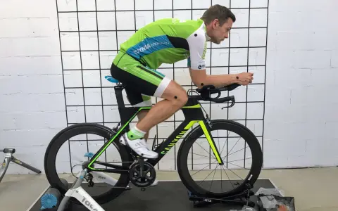 Mężczyzna w stroju kolarskim podczas profesjonalnego fittingu roweru. Czarny rower z zielonymi akcentami stoi na trenażerze.