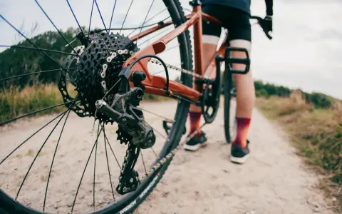 Zbliżenie na tylną przerzutkę roweru gravelowego, gdzie widać sprzęgło w przerzutce. Rower stoi na szutrowej drodze, a w tle widać sylwetkę rowerzysty.