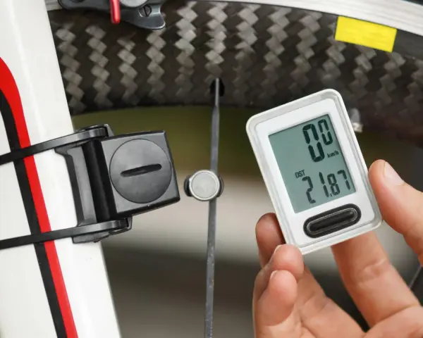 Dłoń trzyma licznik rowerowy pokazujący dystans 21.87 km. Obok widoczny element czujnika na obwodzie koła rowerowego.
