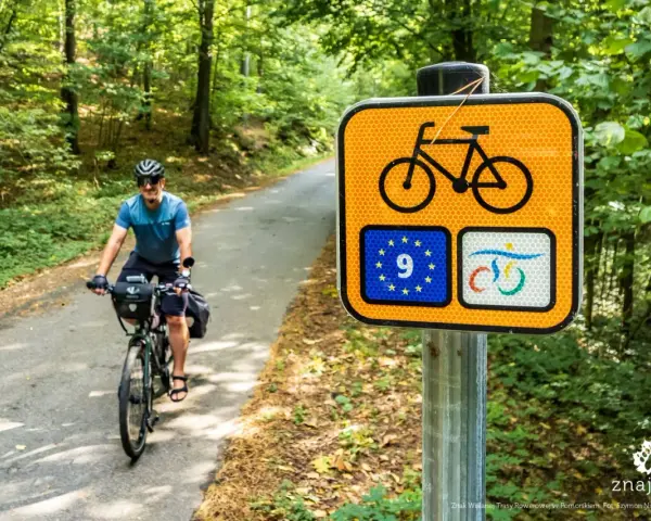 Rowerzysta na leśnej drodze, obok znak szlaku rowerowego krajowego z symbolem UE.