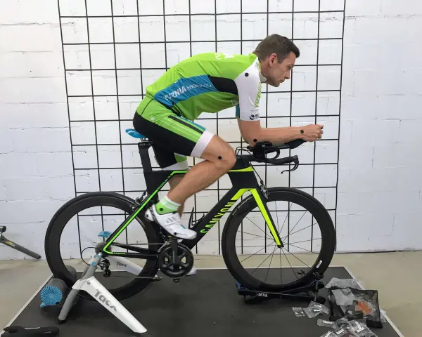 Mężczyzna w stroju kolarskim podczas profesjonalnego fittingu roweru. Czarny rower z zielonymi akcentami stoi na trenażerze.
