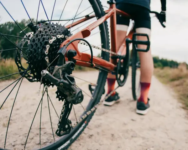 Zbliżenie na tylną przerzutkę roweru gravelowego, gdzie widać sprzęgło w przerzutce. Rower stoi na szutrowej drodze, a w tle widać sylwetkę rowerzysty.