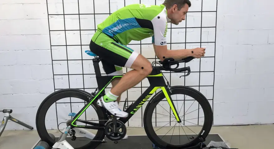 Mężczyzna w stroju kolarskim podczas profesjonalnego fittingu roweru. Czarny rower z zielonymi akcentami stoi na trenażerze.