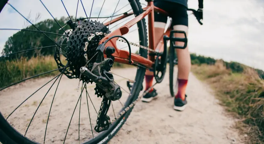 Zbliżenie na tylną przerzutkę roweru gravelowego, gdzie widać sprzęgło w przerzutce. Rower stoi na szutrowej drodze, a w tle widać sylwetkę rowerzysty.