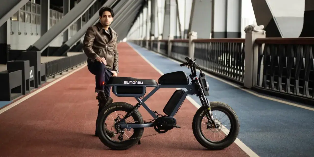 Młody mężczyzna w skórzanej kurtce opiera się o stylowy rower elektryczny. Ten model, z imponującym zasięgiem 300 km, idealnie sprawdzi się na długie wyprawy.