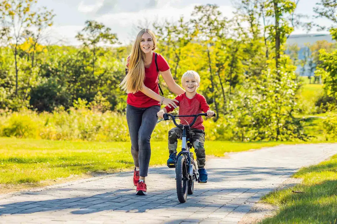 Mama pomaga synkowi, kt&oacute;ry ma 10 latek i nie umie jeszcze jeździć na rowerze. Uśmiechnięci, na ścieżce w parku.