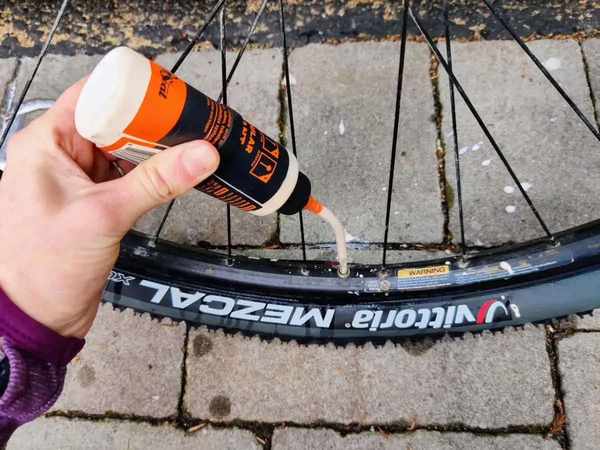 Ręka wlewa płyn do opony tubeless przez wentyl. Widać butelkę z pomarańczowo-czarną etykietą i napis 