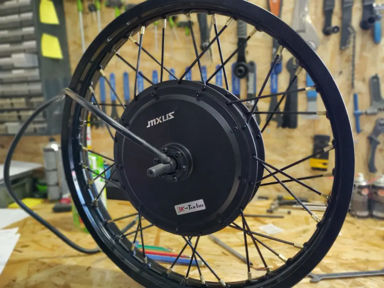 Koło rowerowe z silnikiem MXUS 3K-Turbo w trakcie wymiany piasty. Narzędzia widoczne w tle sugerują prace warsztatowe.