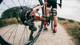 Zbliżenie na tylną przerzutkę roweru gravelowego, gdzie widać sprzęgło w przerzutce. Rower stoi na szutrowej drodze, a w tle widać sylwetkę rowerzysty.